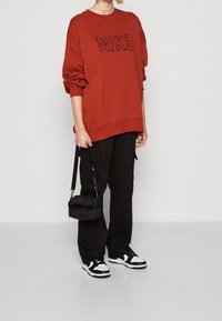 Person som bär en röd Nike-tröja, svarta cargo-byxor, svart-vita sneakers och håller en liten svart handväska vid sin sida.