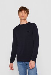Pulover tricotat bleu marin, cu guler rotund și manșete cu model cu nervuri, având un mic logo alb pe piept. Purtați-l cu blugi albaștri deschiși.