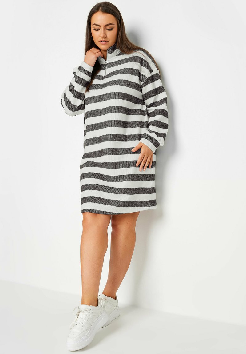 Randig klänning med hög krage, prydd med svarta och vita horisontella ränder, tillverkad av ett mjukt, texturerat tyg och slutar ovanför knäet.