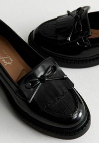 Mocassins en cuir synthétique noir avec un frange texturé et un détail en nœud. Bout rond et semelle en caoutchouc avec un motif de tread subtil.