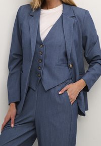 Skrojony na miarę garnitur trzyczęściowy w średnim niebieskim kolorze, z dopasowaną kamizelką zapinaną na pięć guzików, kołnierzem z klapami oraz bocznymi kieszeniami na marynarce.