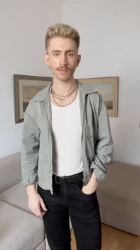 Veste gris clair sur une chemise blanche, pantalon noir et colliers en argent superposés. L'arrière-plan présente des œuvres d'art encadrées et un canapé rayé.