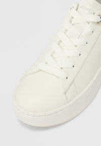 Λευκό δερμάτινο αθλητικό παπούτσι με λευκά κορδόνια, στρογγυλή μύτη και ορατό ασημί οπές, σε απλό ανοιχτό φόντο.