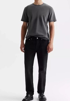 Mann trägt ein ausgewaschenes graues T-Shirt mit kurzen Ärmeln, schwarze Jeans und schwarze Lederschuhe, steht vor einem schlichten weißen Hintergrund.