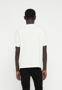 Λευκό κοντομάνικο μπλουζάκι από βαμβάκι, με στρογγυλή λαιμόκοψη και ίσια κοπή· φορεμένο με μαύρο τζιν, εμφανιζόμενο από πίσω.