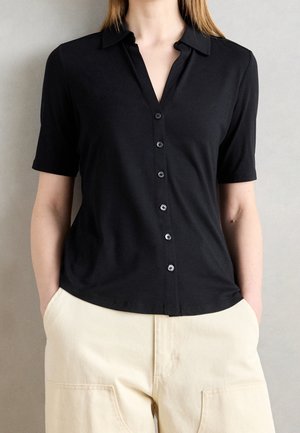 Osoba ubrana w dopasowaną czarną koszulę z krótkim rękawem i kołnierzykiem, wpuszczoną w kremowe spodnie z wysokim stanem i dużymi przednimi kieszeniami.