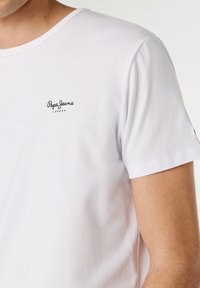 Férfi egyszerű fehér rövid ujjú pólóban, bal mellrészen kis fekete "Pepe Jeans London" logóval.