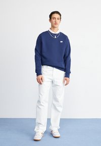 Mörkblå sweatshirt med ribbad manschetter och krage, vita byxor och vita skor med orangea accenter. Modellen står mot en enfärgad bakgrund.
