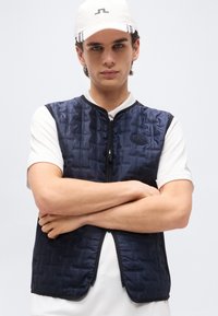 Νεαρός άνδρας με σταυρωμένα τα χέρια φοράει λευκό καπέλο, λευκό κοντομάνικο πουκάμισο και ναυτικό μπλε καπιτονέ αμάνικο γιλέκο με φερμουάρ.