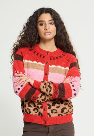 Femme aux longs cheveux bouclés portant un cardigan coloré boutonné avec des motifs rouges, roses, marron et léopard, debout les bras croisés.