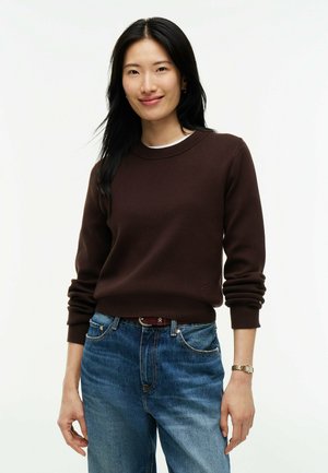 Junge Frau mit langen schwarzen Haaren lächelt, trägt einen braunen Pullover, blaue Jeans, eine Uhr und einen braunen Gürtel vor weißem Hintergrund.