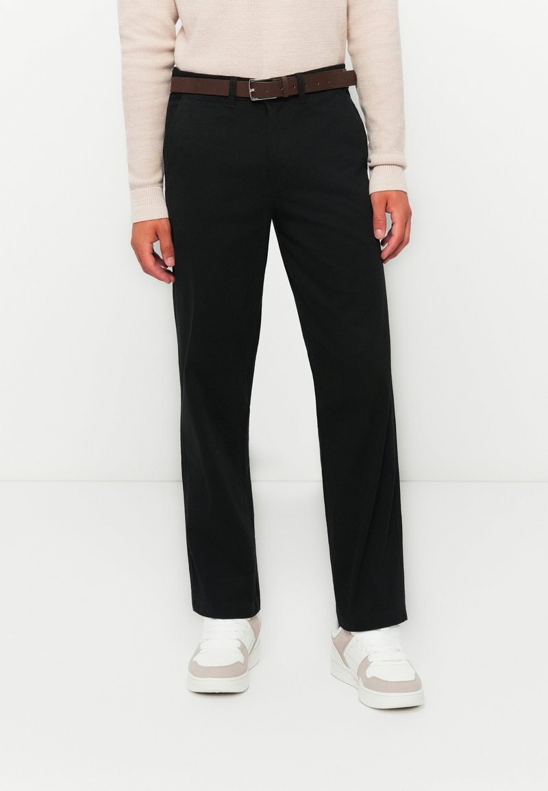 Person wearing black straight-leg pants, brown belt, light beige long-sleeve top, and white sneakers against a white background.