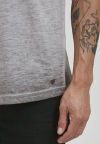 Grå, kortermet skjorte med en myk, teksturert finish. Har en liten logo nederst og kontraststing ved bunnen.