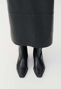 Schwarze Lederstiefel mit hohem Schaft und quadratischer Zehenform sowie seitlichem Reißverschluss. Glatte Textur und minimales Zubehör; kombiniert mit einem schwarzen Lederrock.