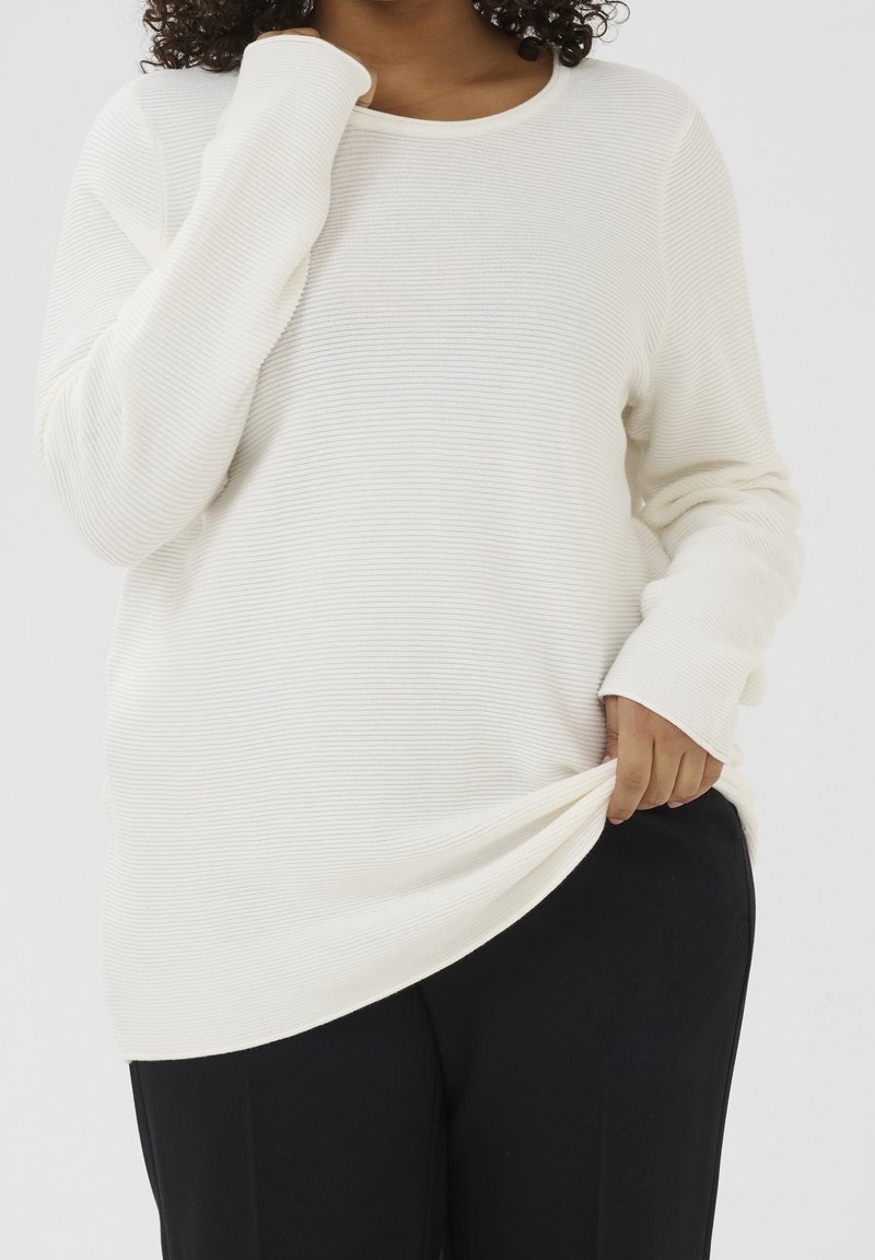 Personne portant une chemise blanche à manches longues côtelée et un pantalon noir, tenant l'ourlet de la chemise d'une main et touchant l'encolure de l'autre.