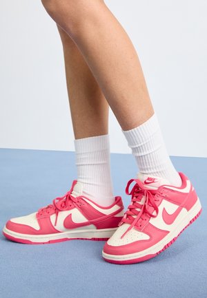 Benen met witte crew-sokken en roze-witte Nike sneakers, staand op een lichtblauwe vloer tegen een effen witte achtergrond.