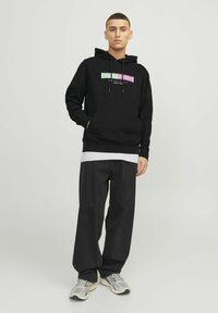 Zwarte hoodie met een voorzak en trekkoorden, met een kleurrijke grafische logo. Gecombineerd met zwarte wijde broek en sneakers.