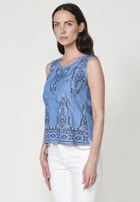Top senza maniche in denim blu con motivi geometrici ricamati, collo rotondo e orlo leggermente accorciato. Abbinato a pantaloni bianchi.