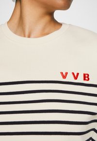 Personne portant un pull crème avec des rayures horizontales noires et les lettres VVB brodées en rouge sur la poitrine.