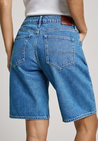 Persona vestida con pantalones cortos de mezclilla de color azul claro hasta la rodilla y una blusa blanca sin mangas, de pie con las manos en los bolsillos, vista trasera.
