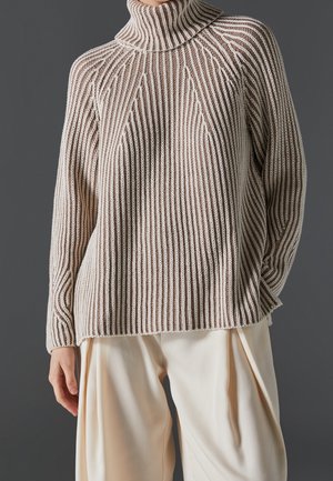 Pull à col roulé beige et blanc à rayures étroitement côtelé, associé à un pantalon ample blanc cassé sur fond gris.