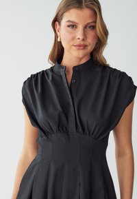 Crna haljina s mandarin ovratnikom, kratkim puf rukavima, zatvaranjem na dugmad i nabranim strukom. Glatka tkanina s elegantnim završetkom.