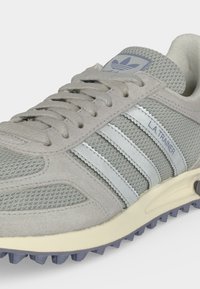 Γκρι αθλητικό παπούτσι Adidas L.A. Trainer με άνω μέρος από δίχτυ και σουέτ, λευκές ρίγες, υφασμένο πέλμα και ετικέτα με λογότυπο στη γλώσσα.