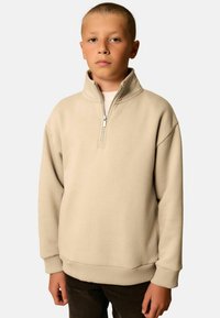 Junge mit kurzen Haaren, der einen beigen Pullover mit Viertelreißverschluss und dunklen Hosen trägt, steht vor einem schlichten hellen Hintergrund.