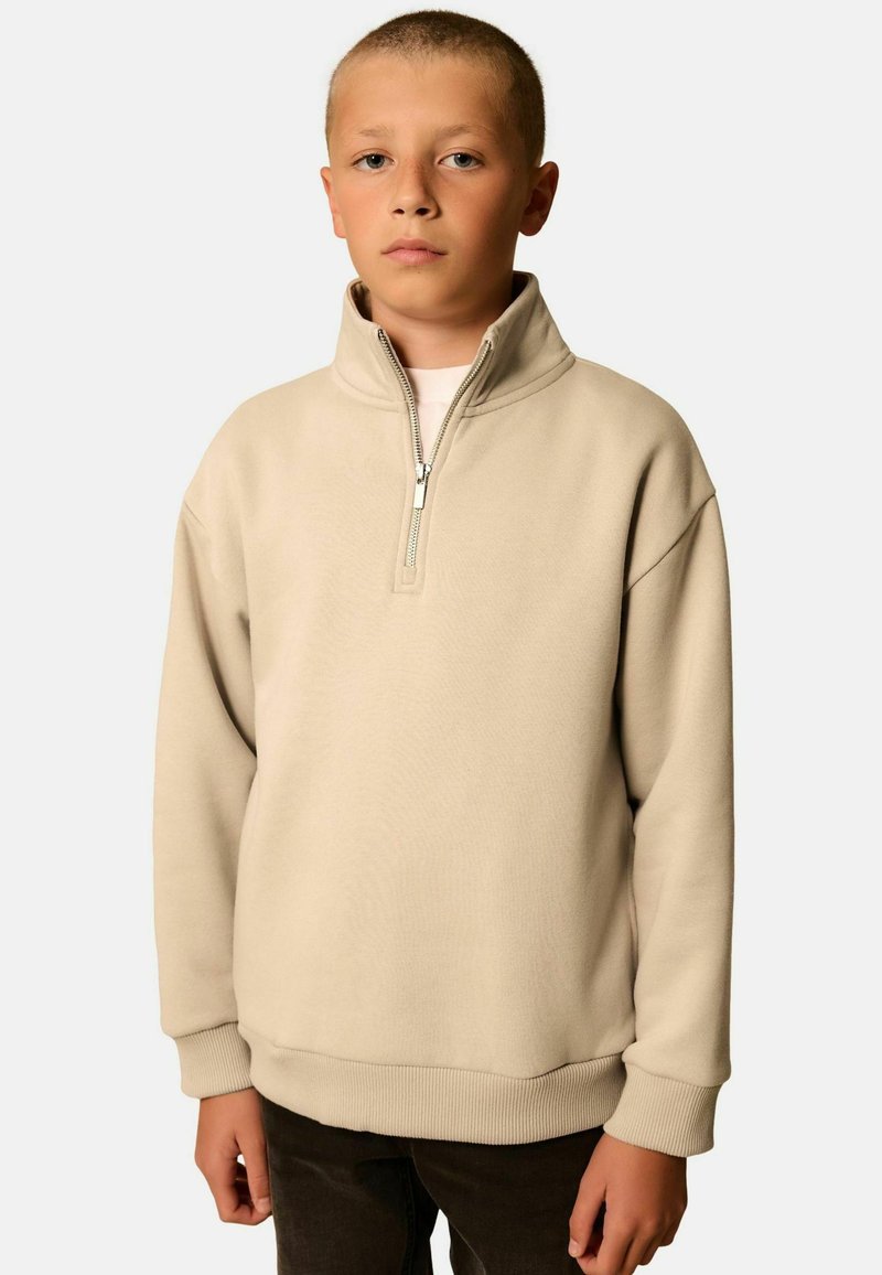 Junge mit kurzen Haaren, der einen beigen Pullover mit Viertelreißverschluss und dunklen Hosen trägt, steht vor einem schlichten hellen Hintergrund.
