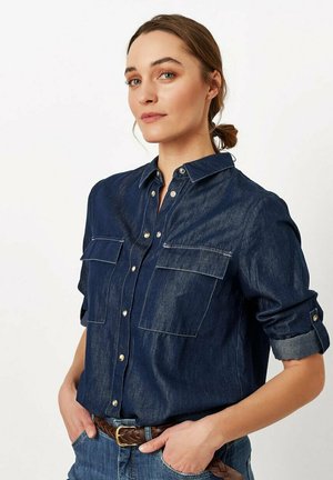 Denim shirt in dark blue, featuring two chest pockets, button-up front, and rolled sleeves. Smooth texture with contrasting stitching.