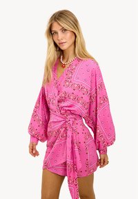 Robe rose à motifs avec manches longues, présentant un design portefeuille et une taille nouée. Le tissu a une texture douce avec des accents floraux.