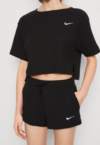 Woman wearing black ribbed Nike short-sleeve crop top and matching shorts with white Nike swoosh logos, hand in pocket, light skin tone.