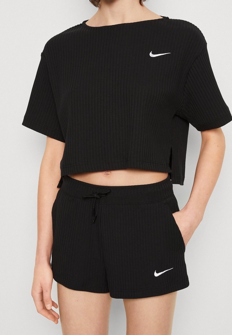 Woman wearing black ribbed Nike short-sleeve crop top and matching shorts with white Nike swoosh logos, hand in pocket, light skin tone.