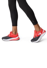 Scarpe atletiche con tomaia in rete grigia e accenti corallo brillante, caratterizzate da una suola bianca con una base rosa e un motivo di battistrada nero.
