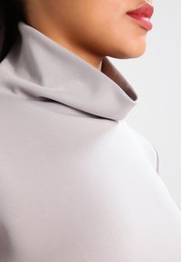 Ljusgrå höghalstern med mjukt, stretchigt tyg. Har en vikt krage och en åtsittande design som framhäver en slank silhuett.