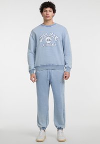 Šviesiai mėlynas džemperis su baltu užrašu "Guess Jeans California", derinamas su atitinkamomis kelnėmis, ant kurių yra logotipas; abi dalys turi raibuoto kraštą.