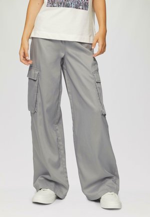 Grijze cargobroek met wijde pijpen, voorzien van zijzakken en een soepele textuur. Gepaard met witte sneakers en een lichtgekleurde T-shirt.