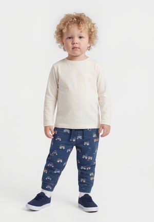 Bambino con capelli biondi ricci che indossa una maglietta a maniche lunghe color crema, pantaloni blu con stampe di trattori, calzini bianchi e scarpe da ginnastica blu scuro, in piedi su sfondo bianco.