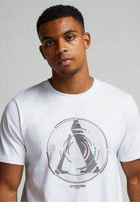 Valkoinen puuvillateeppari, jossa on pyöreä grafiikka mustista ja vaaleanpunaisista viivoista. Suunnittelu luo 3D-efektin, keskellä teksti "ALESSANDRO."