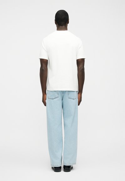 Άνδρας που φοράει απλό λευκό κοντομάνικο πουκάμισο, ανοιχτά μπλε τζιν με φαρδιά μπατζάκια και μαύρα παπούτσια, στέκεται γυρισμένος με την πλάτη προς έναν λευκό φόντο.