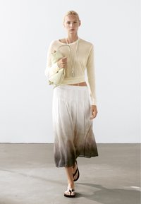 Lichtbeige gebreide trui met lange mouwen, gecombineerd met een vloeiende rok tot de enkels met een ombre-gradiënt van wit naar donkerbruin. Zwarte sandalen.