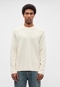 Krēmkrāsas garroku džemperis ar apaļu kakla izgriezumu, fakturētu audumu, plakaniem šuvumiem un vaļēju piegriezumu, kombinēts ar zilām džinsiem ar klasisku piegriezumu.