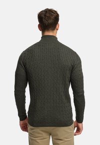 Mörkgrön kabelstickad tröja med hög krage och långa ärmar. Ribbad struktur med figurnära silhuett.