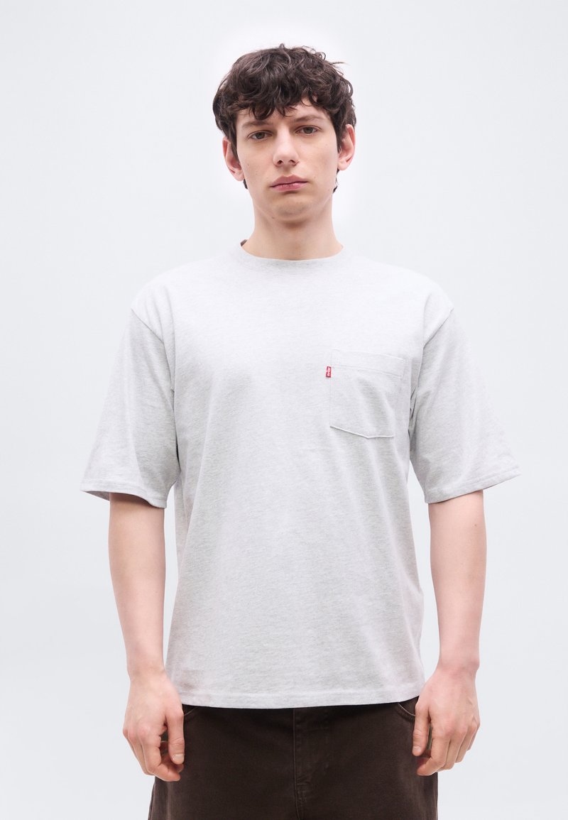 Giovane uomo con capelli ricci e scuri che indossa una maglietta a maniche corte grigio chiaro con una tasca sul petto e pantaloni scuri, su uno sfondo semplice.