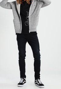 Sudadera gris con cremallera, camiseta gráfica negra con diseño de gato, jeans oscuros ajustados y zapatillas Vans negras con cordones y suelas blancas.