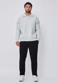 Pull gris clair en maille avec col, associé à un pantalon noir. Présente une coupe décontractée avec des poignets et un ourlet côtelés. Le modèle est debout.