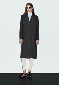 Cappotto grigio in misto lana, doppiopetto, con grandi rever e tasche laterali, abbinato a leggings color crema e sandali neri. Il modello indossa occhiali da sole verdi.