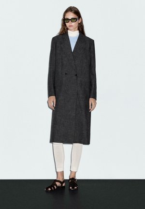 Manteau double boutonnage en laine mélangée gris, avec de grands revers et des poches latérales, associé à un legging crème et des sandales noires. Le mannequin porte des lunettes de soleil vertes.