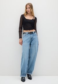 Top corto a maniche lunghe in pizzo nero abbinato a jeans a gamba larga azzurri e cintura nera. La modella indossa stivaletti neri.