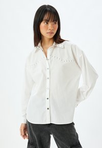 Camicia bianca con bottoni automatici, borchie decorative lungo il collo e maniche lunghe dal fit morbido. Tessuto liscio e leggero.